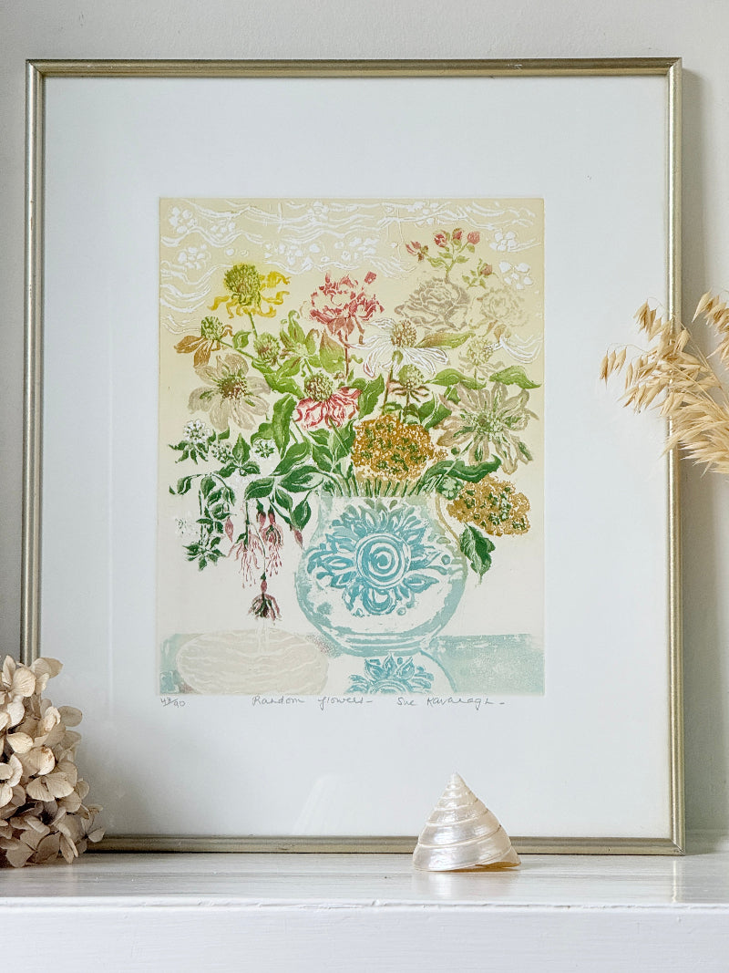 A framed botanical art print featuring a blue patterned vase filled with colorful flowers, displayed on a white surface with decorative dried grass and a shell nearby.
