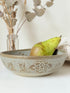 Studio pottery bowl with carved floral and geometric decoration in neutral glaze, photographed on a white surface with a pear inside.