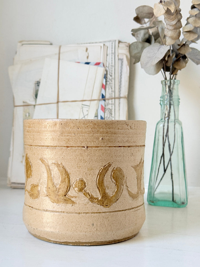 A handmade beige ceramic pot with gold Arabic calligraphy patterns, styled on a white surface next to a glass bottle vase with dried eucalyptus and a stack of vintage letters.