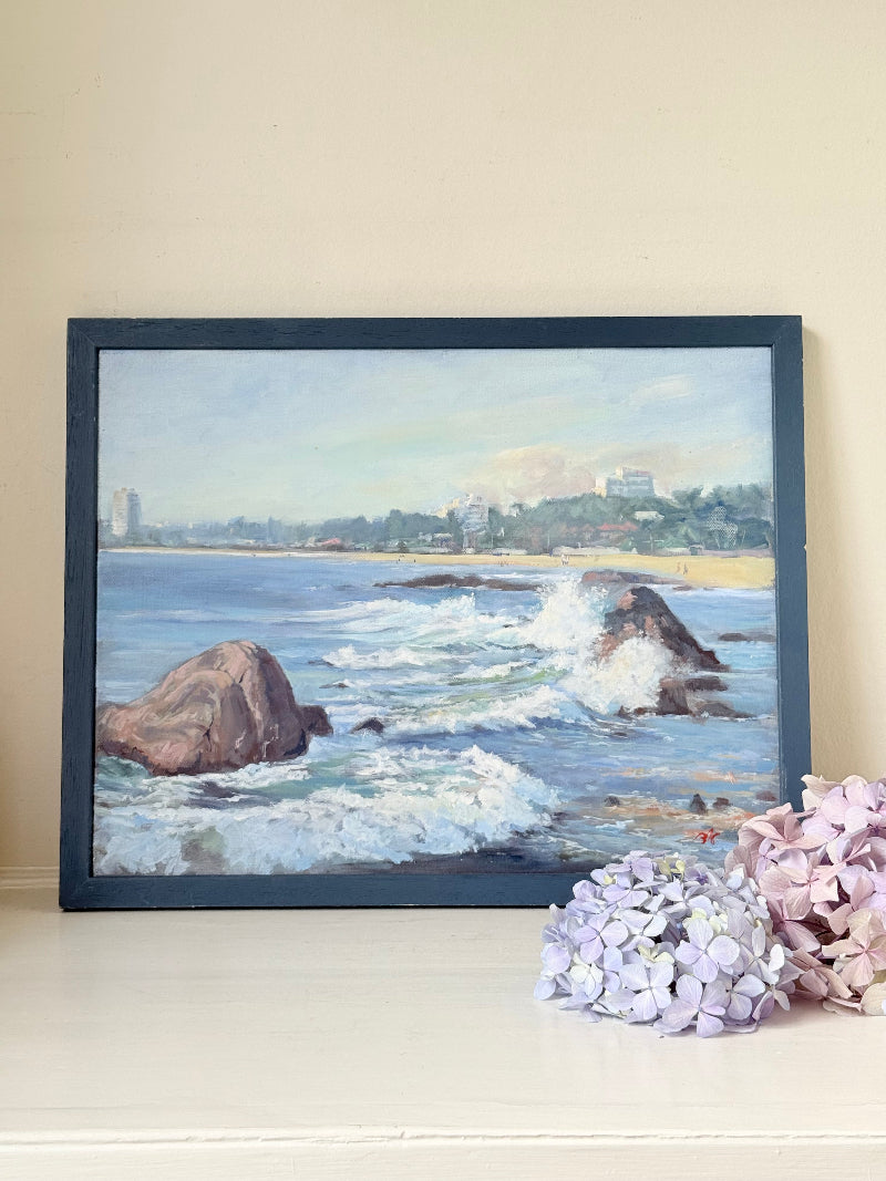 Original oil painting of waves crashing against rocks, signed seascape in slate blue wooden frame on white surface