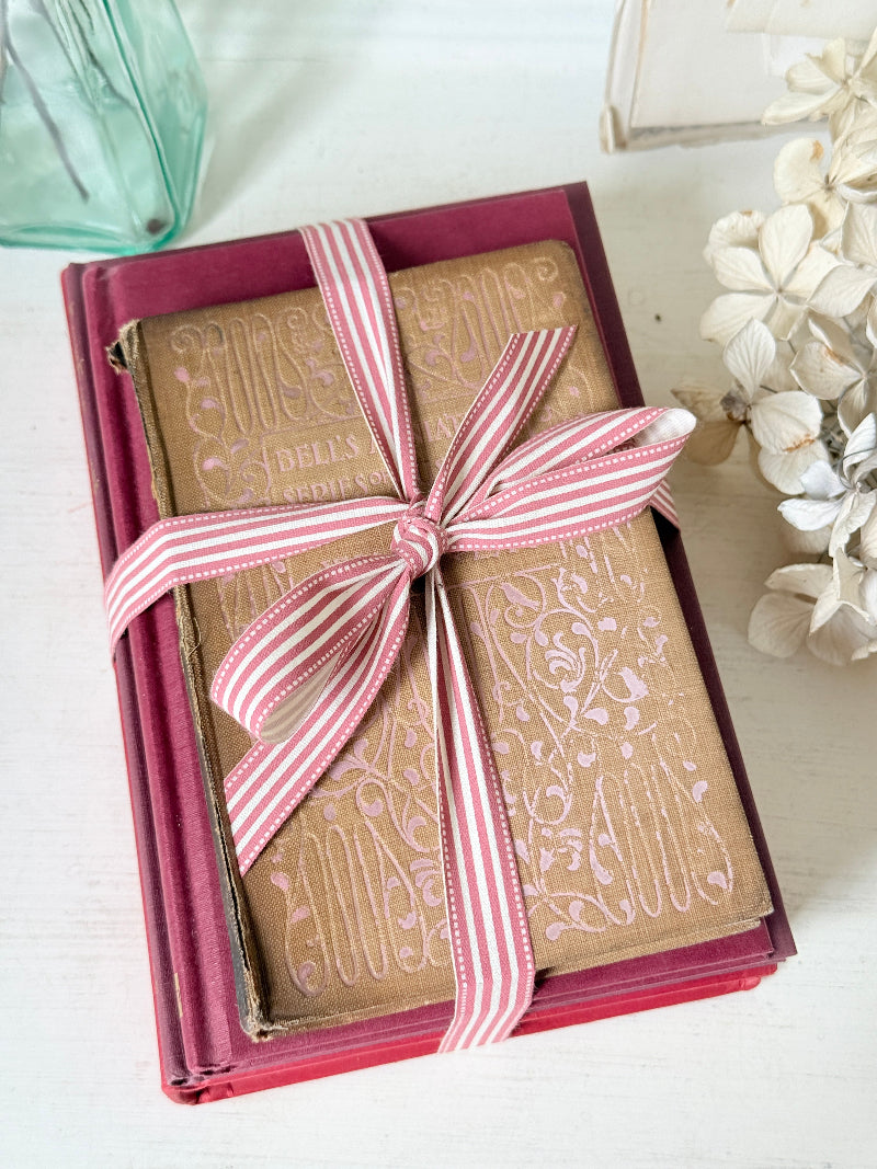 Stack of cloth bound vintage books in red and warm pink tones tied with striped ribbon and dried flowers