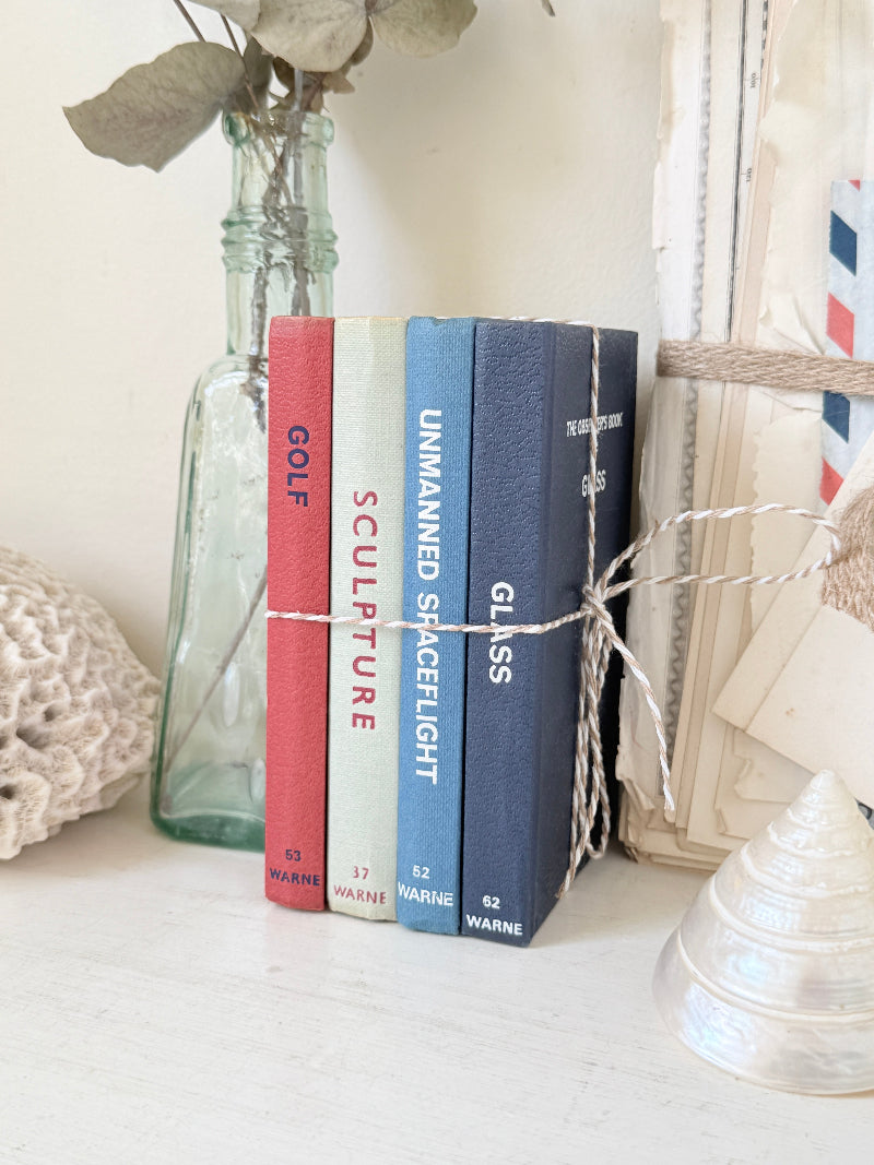 Four decorative book spines tied together with twine, standing upright on a white surface beside a glass bottle, a crochet bag, and a framed print. The spines are colored red, off-white, light blue, and dark navy, with text reading GOLF, SCULPTURE, UNMANNED SHAPELIGHT, and GLASS.