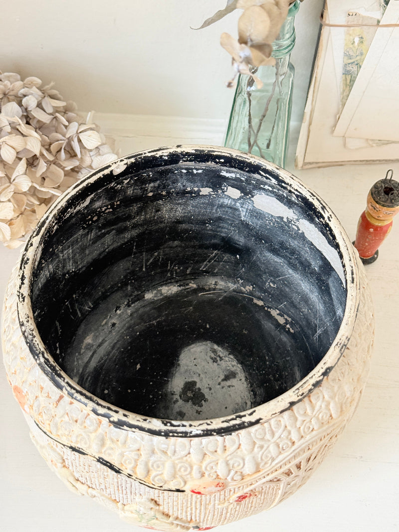 An antique carved ceramic planter pot with a distressed off-white exterior and a dark blackened interior, shown on a white surface with vintage decor in the background.