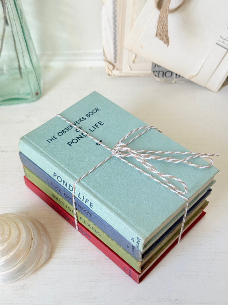 A stack of five vintage-style hardcover books tied with white and pink striped twine, placed on a white tabletop with decorative props in the background.