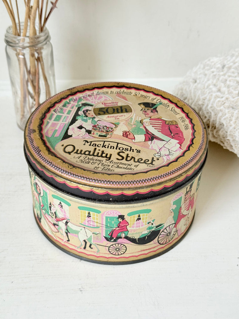 Vintage Quality Street tin from 1986 with colourful illustrated street scenes and pastel design, photographed on a white surface.