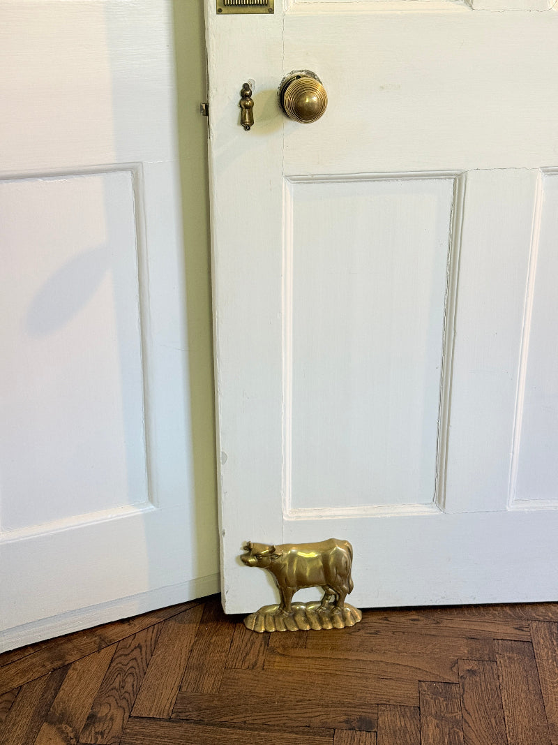 A brass cow door stopper resting on a wooden floor in front of a white paneled door.