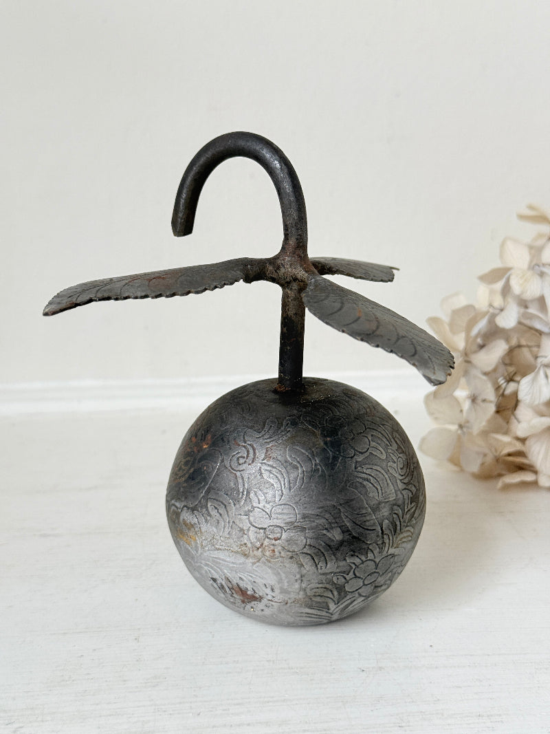 An antique-style metal hanging bell with a round engraved body, dark hook-shaped top, and wing-like side extensions, displayed against a light neutral background with dried flowers at the side.