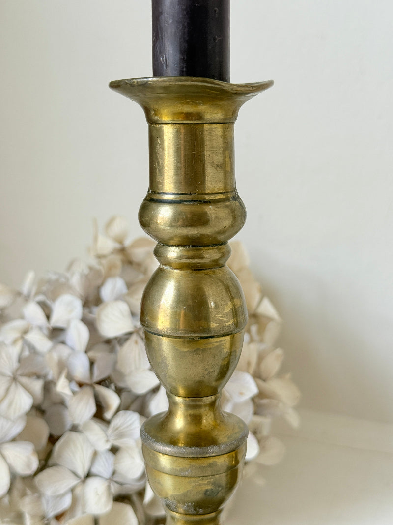 Pair of turned brass candlesticks with square stepped bases and black taper candles on a white surface, styled with dried flowers