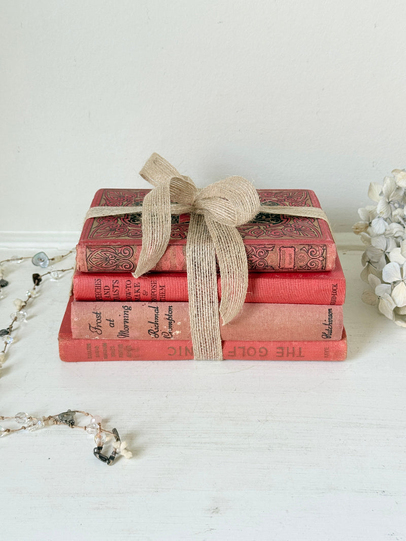 A stack of four vintage-style decorative books in coral red and pink tones, tied with a natural jute ribbon bow, displayed on a white surface against a white wall.