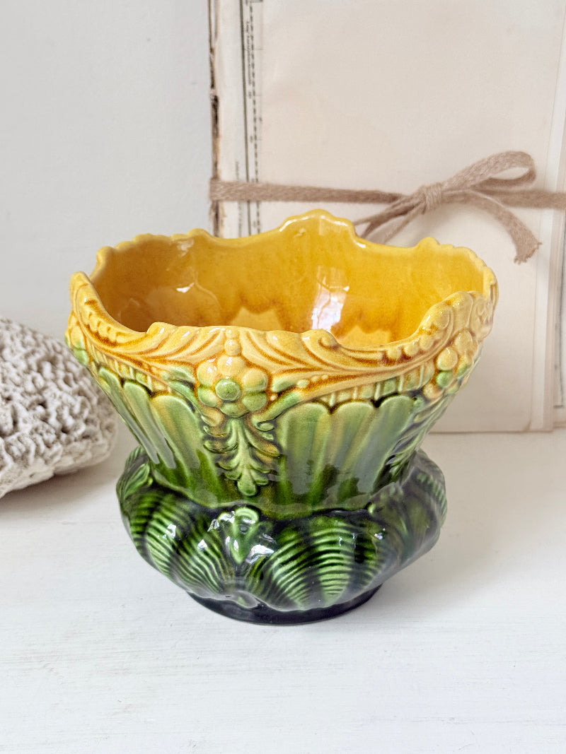 A decorative ceramic planter pot with a yellow and green glaze, featuring a scalloped rim, embossed floral details, and a ribbed base, set against a rustic white background.