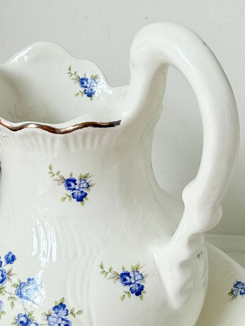 A white ceramic pitcher with a decorative handle, featuring blue floral patterns and a metallic gold rim, shown against a neutral light gray background.