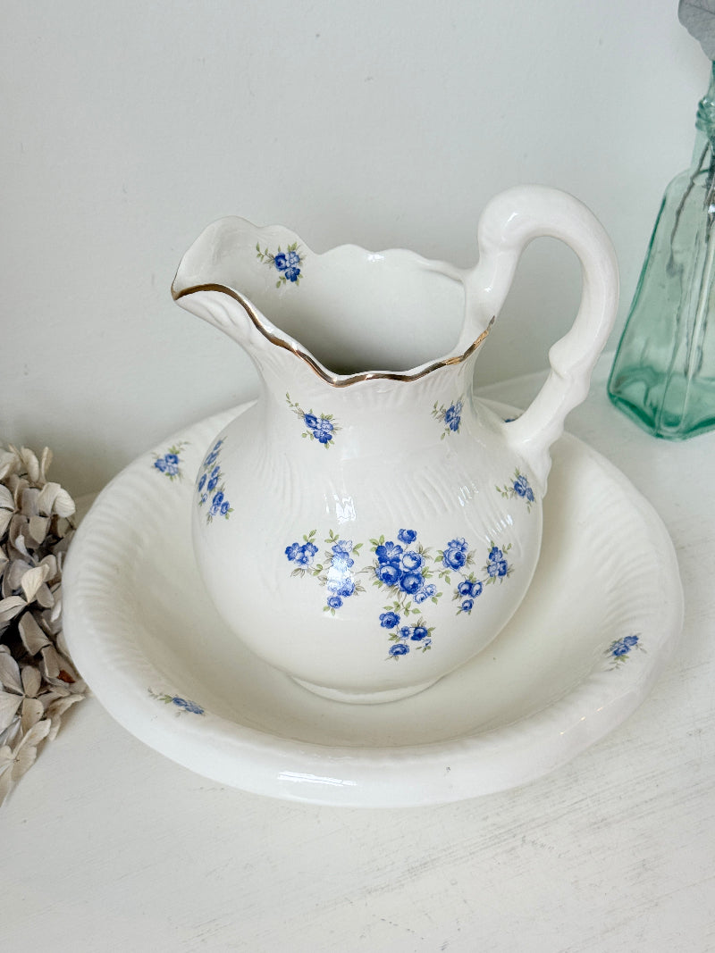 A white ceramic pitcher with a matching saucer, decorated with blue floral patterns and a thin gold rim, set against a light background with decorative props.