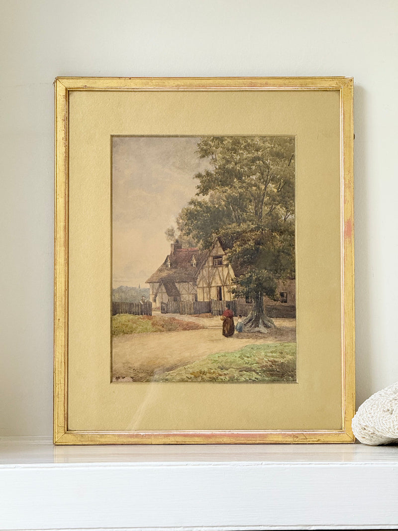 Antique framed watercolour of English village street with thatched cottages and figures, in gilt frame with wide mount, on white surface.