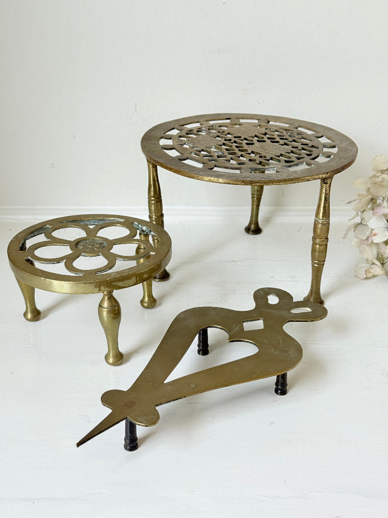 Vintage brass trivet trio with ornate cutwork designs and aged patina, photographed on a white surface