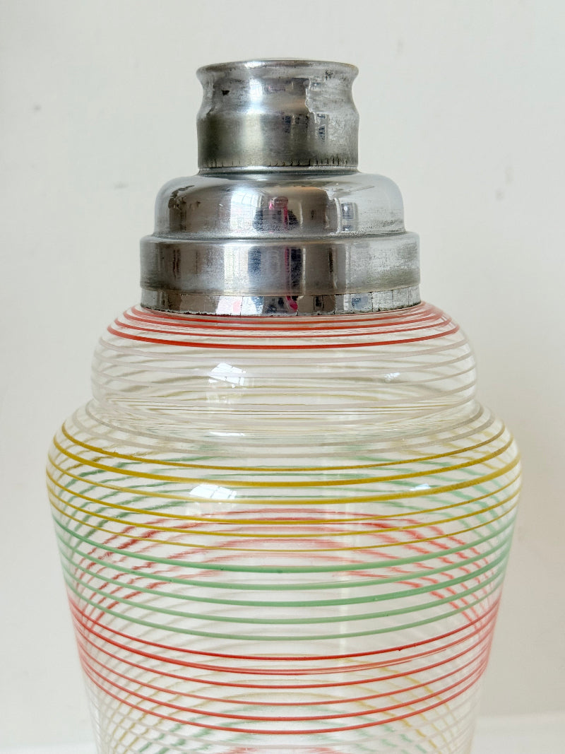 Vintage glass cocktail shaker with multicoloured stripes and silver lid photographed on a white surface.