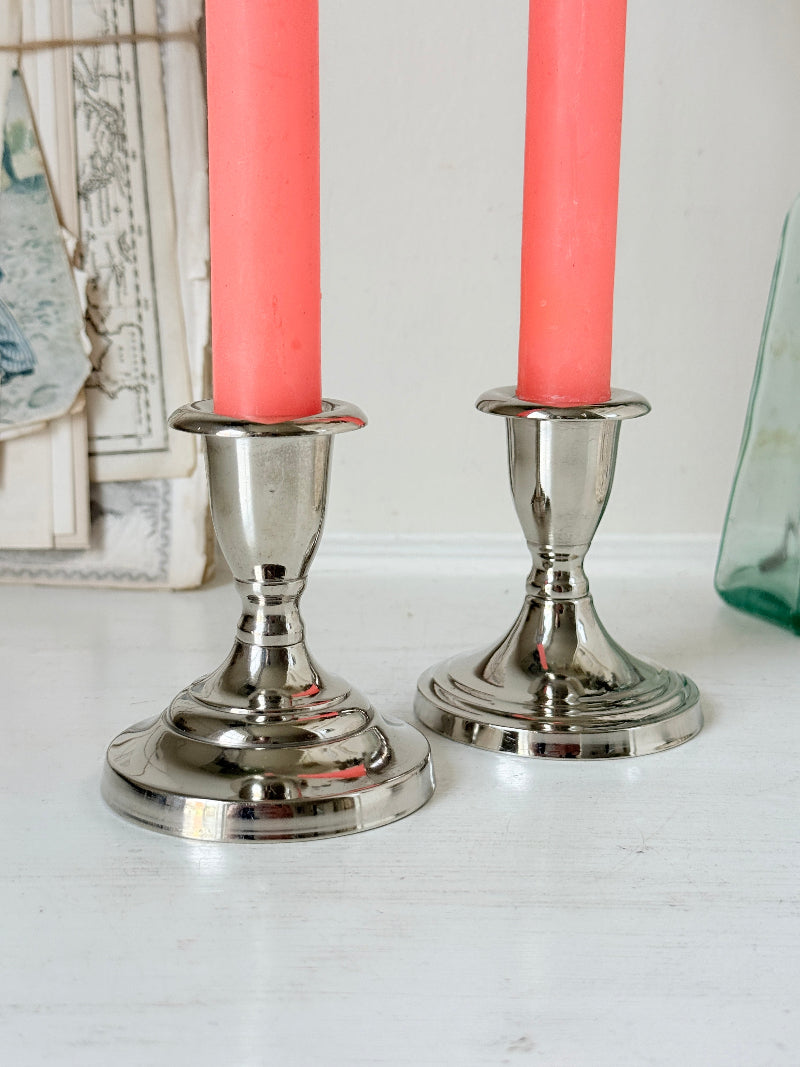 Pair of silver tone candlesticks with urn sconces and round stepped bases holding coral candles, photographed on a white surface
