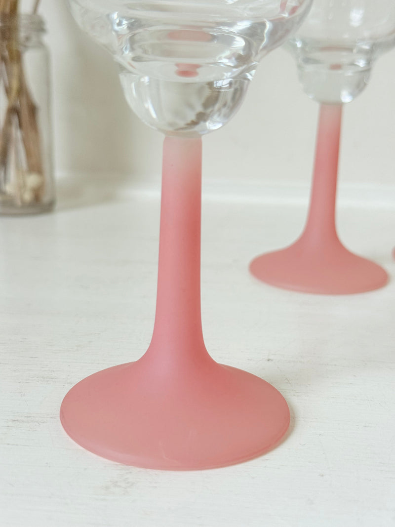 Set of four small vintage wine glasses with clear bowls and pink stems photographed on a white surface.