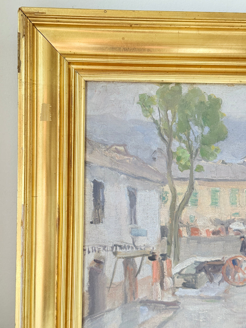 Close-up of a framed painting showing an outdoor street or courtyard scene with a tree, buildings, and figures, surrounded by a wide ornate gold frame.
