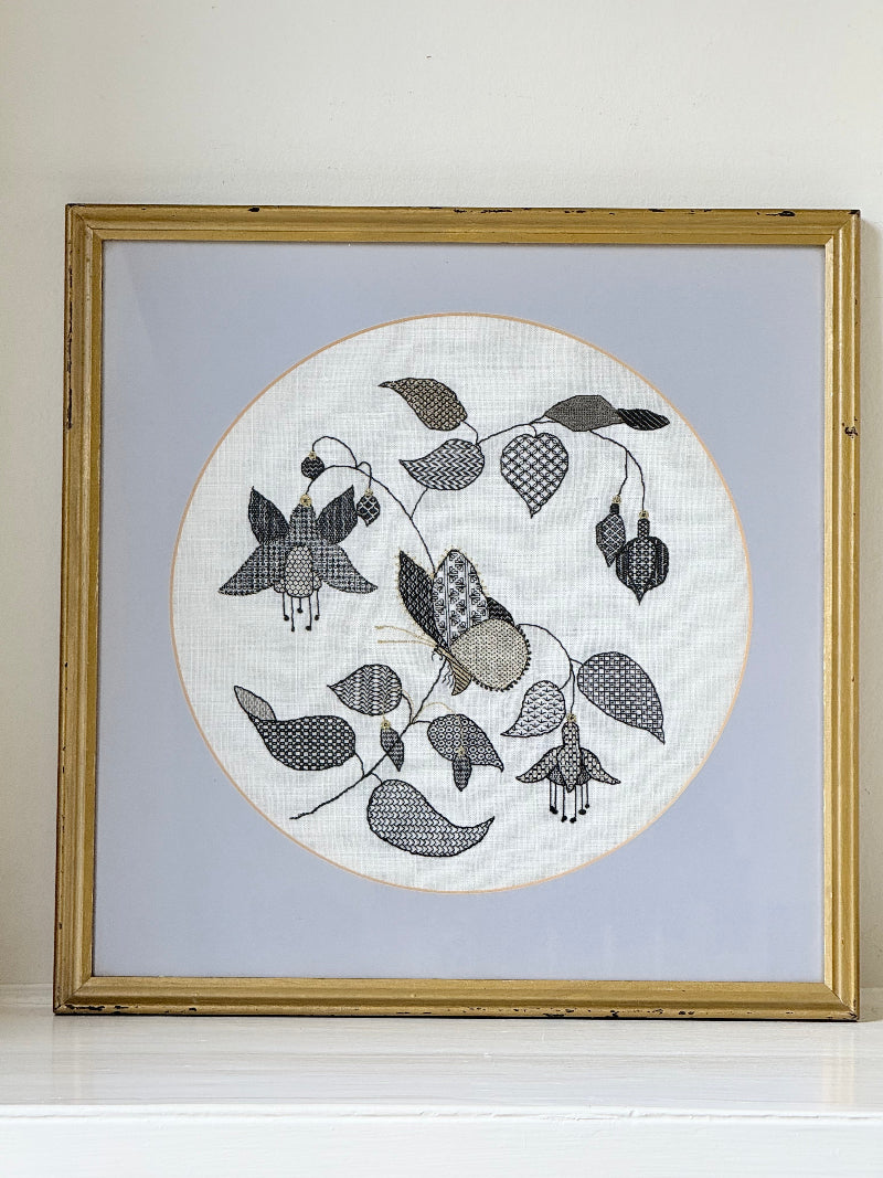 Framed circular hand embroidery of birds and fuchsia flowers in black and grey on white linen, set in gold frame, white surface background.