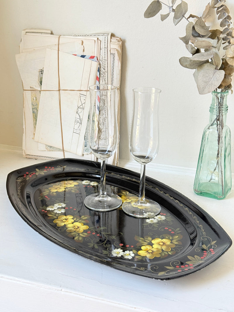 A black oval decorative tray with a floral pattern, styled on a white surface with two clear champagne flutes, a bundle of vintage letters, and a green glass bottle vase with dried leaves.