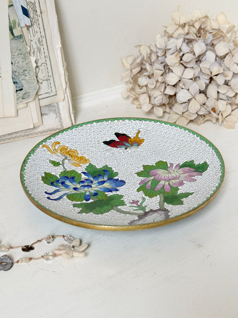 A round decorative ceramic tray with a white textured glaze, featuring hand-painted colorful flowers (blue, pink, yellow), green leaves, a butterfly, and a gold rim, styled on a white surface with dried flowers and a jewelry strand in the background.