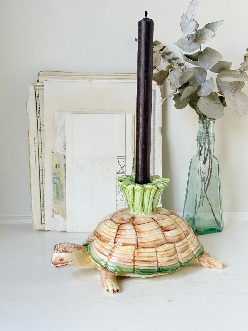 A ceramic turtle-shaped candle holder with a striped tan and green shell, holding a tall black pillar candle, styled with a vintage book and a glass vase on a white surface.