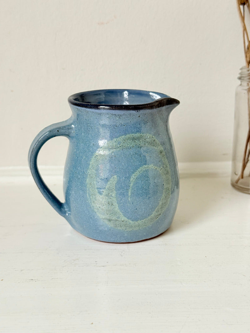 Vintage studio pottery milk jug with blue and green glaze, handmade with a rounded form and sturdy handle, styled on a white surface.