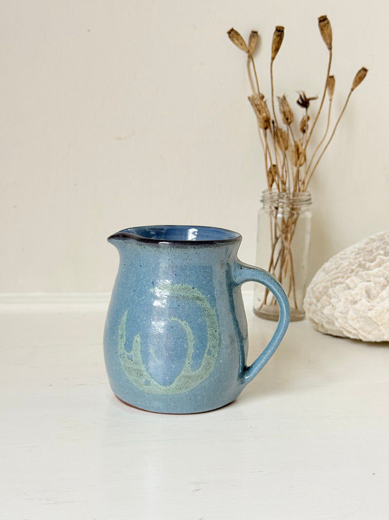 Vintage studio pottery milk jug with blue and green glaze, handmade with a rounded form and sturdy handle, styled on a white surface.