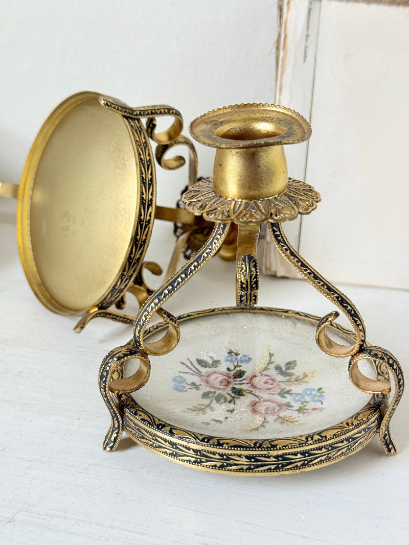 An ornate vintage-style gold and black metal wall sconce with a floral enamel dish and a separate circular mirror or glass panel, shown on a white painted surface.