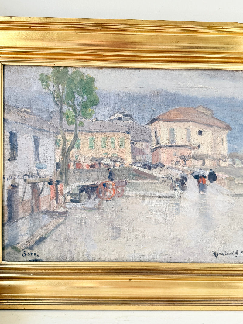 A framed impressionist oil painting depicting a European street scene with buildings, a tree, and figures with umbrellas, housed in a wide gold frame.