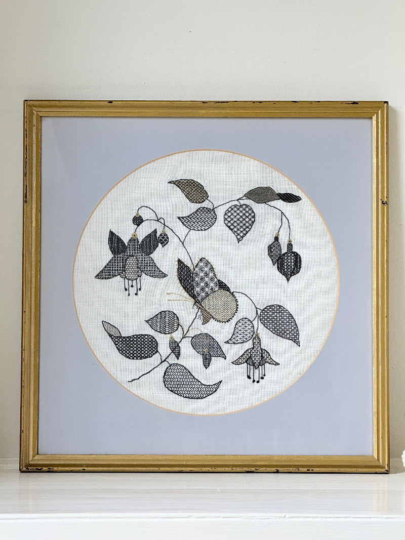 Framed circular hand embroidery of birds and fuchsia flowers in black and grey on white linen, set in gold frame, white surface background.