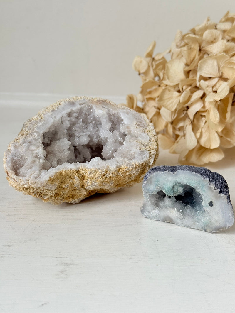 Pair of natural quartz geodes, one white and one lilac-toned, styled on a white surface with dried hydrangea