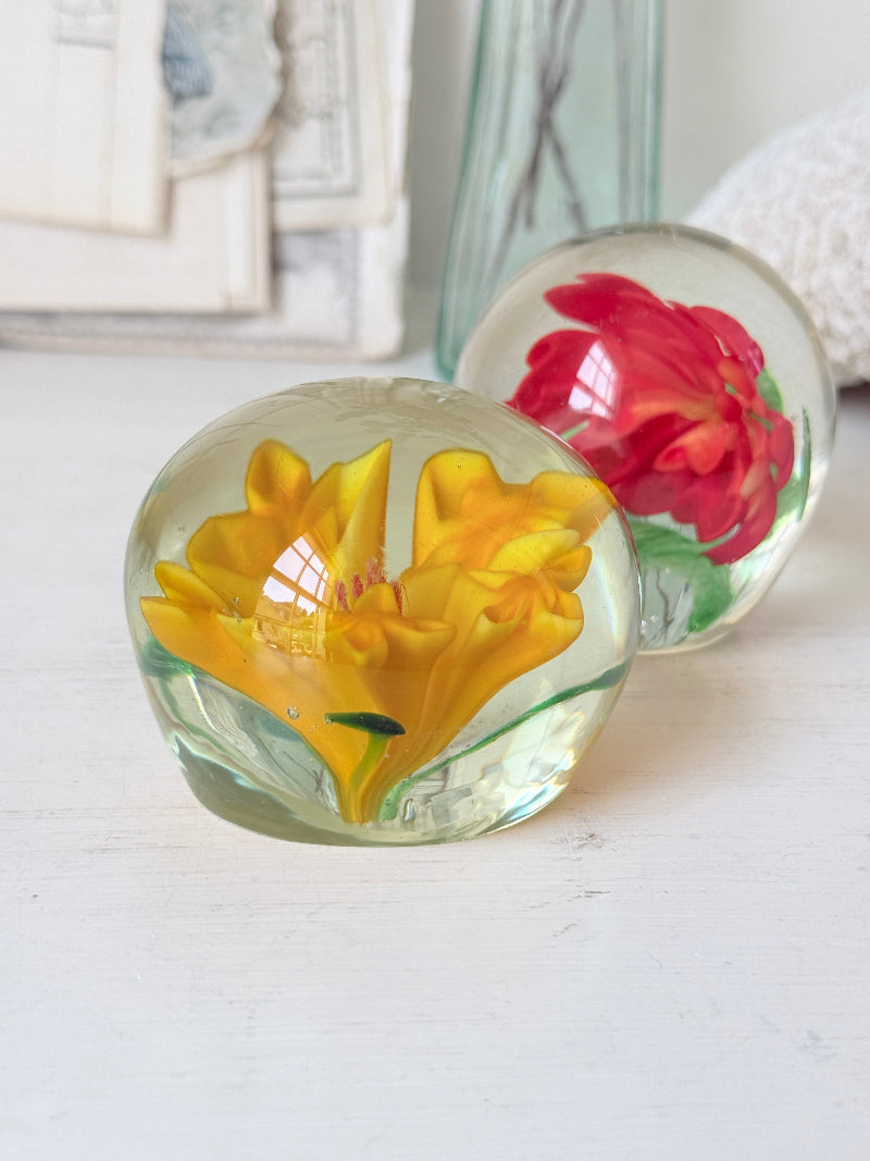 Pair of orange and yellow flower glass paperweights with green leaves, photographed on a white surface