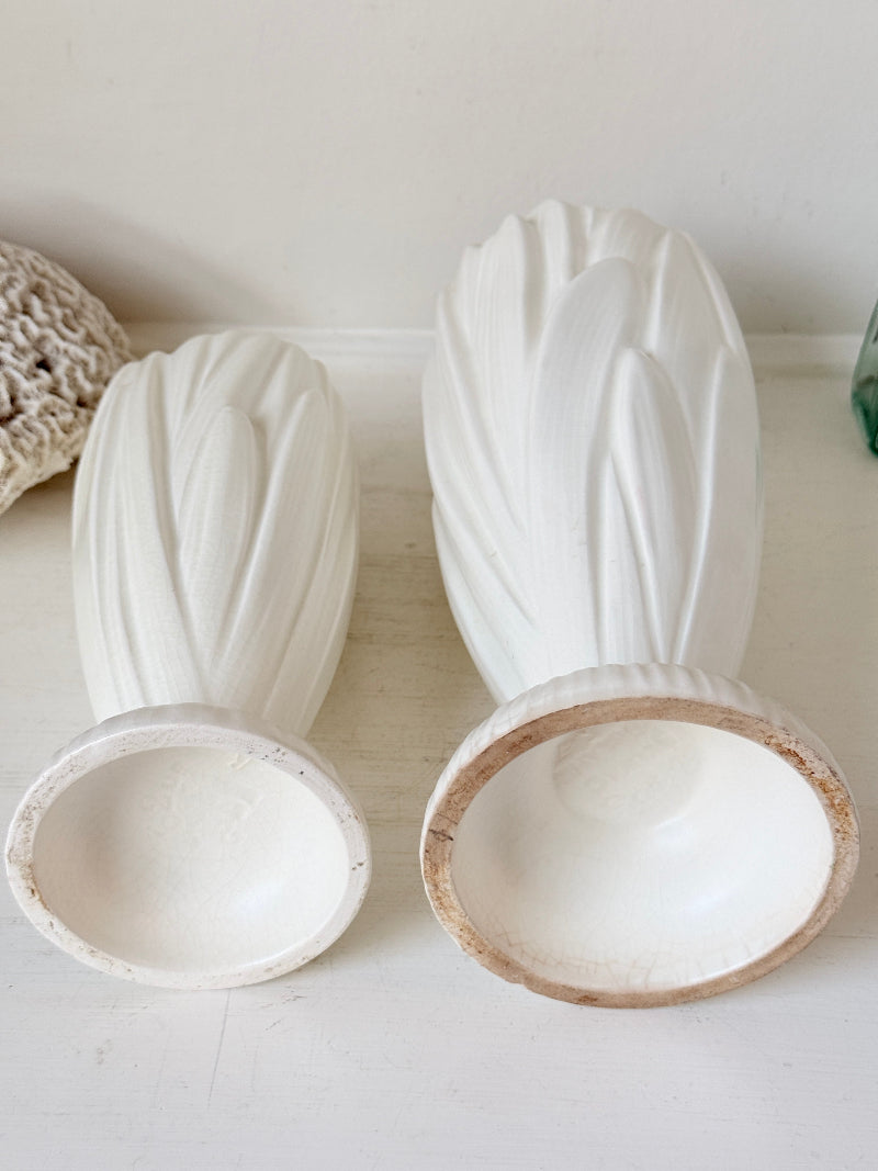 Two white ceramic bud vases with textured leaf-like detailing, shown on a light surface against a white wall, with one vase standing upright and the other upside down to display the interior.