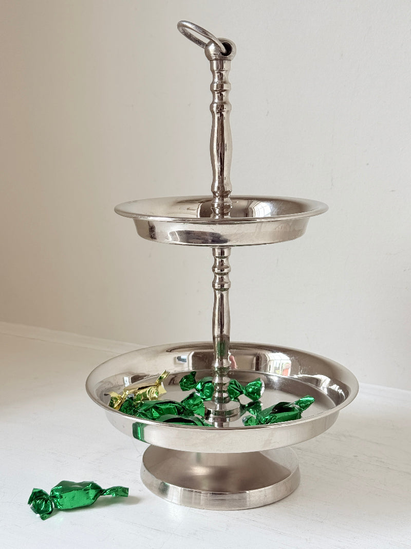 Two tier silver tone cake stand with polished reflective finish styled with green wrapped sweets on a white surface