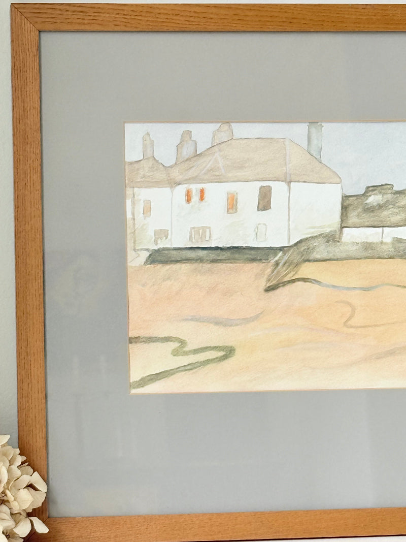 Framed neutral-toned village scene watercolour with figures in light oak-effect frame and pale grey mount, on white surface.