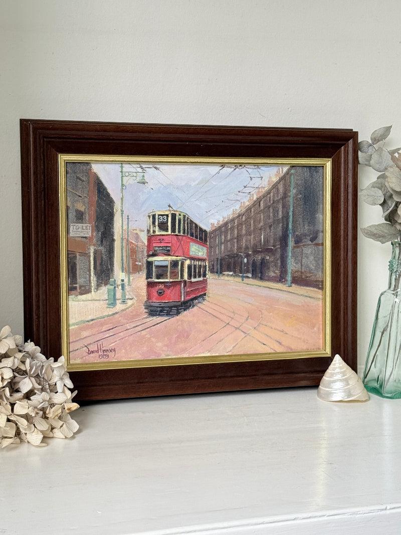 A framed vintage oil painting of a red double-decker tram on a city street, displayed on a white shelf with decorative dried flowers and a glass vase.
