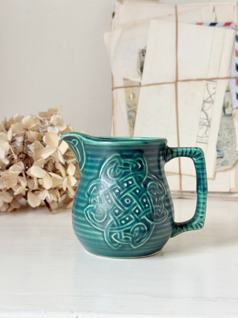 A teal green ceramic pitcher with a raised Celtic knot pattern on the front, set on a white surface with a blurred background of dried flowers and vintage papers.