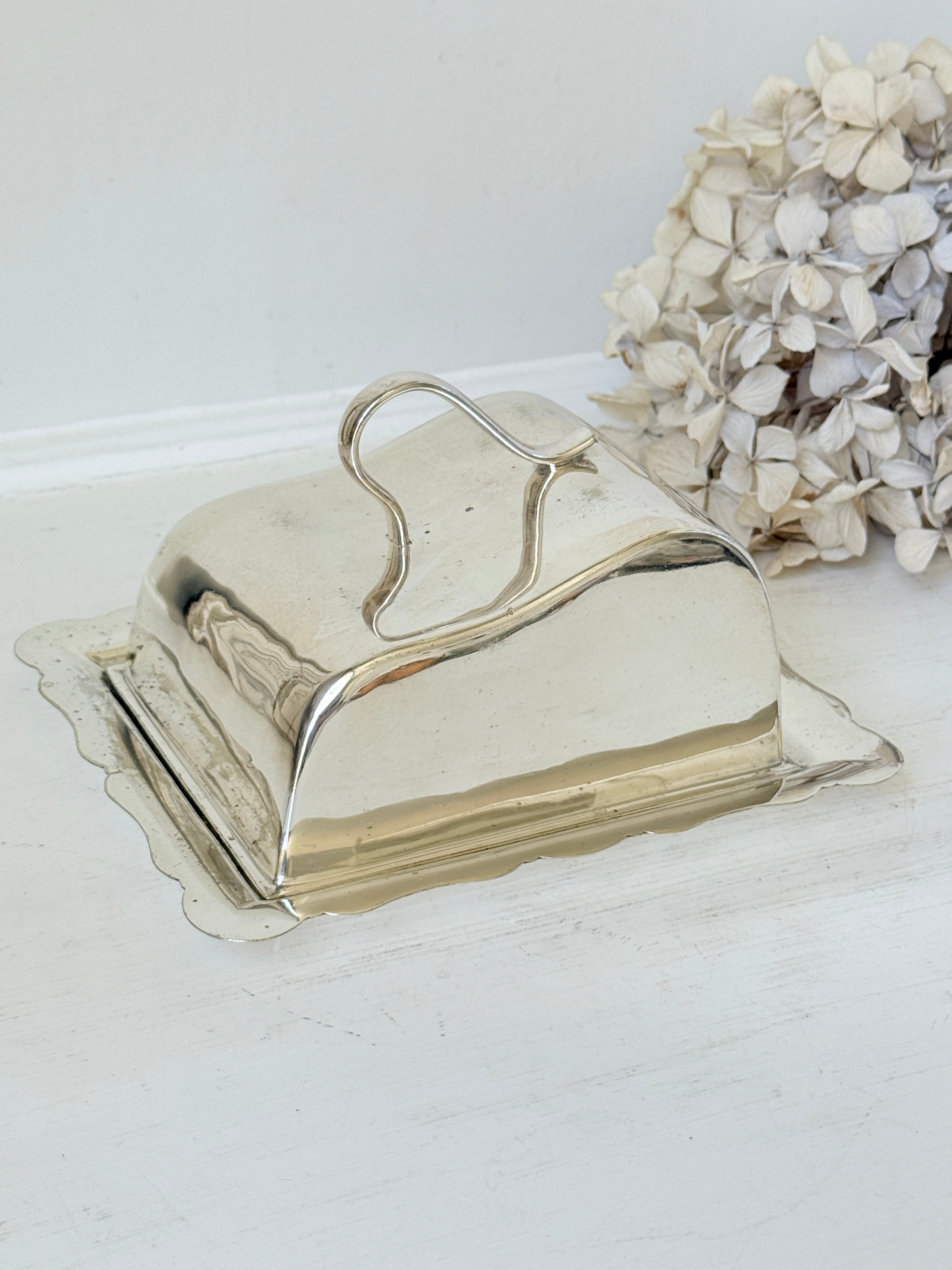 A polished silver butter dish with a hinged lid and scalloped base, shown on a white surface with dried flowers in the background.