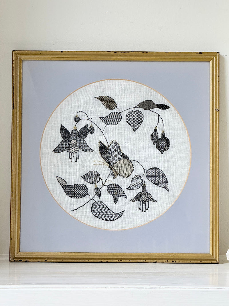 Framed circular hand embroidery of birds and fuchsia flowers in black and grey on white linen, set in gold frame, white surface background.