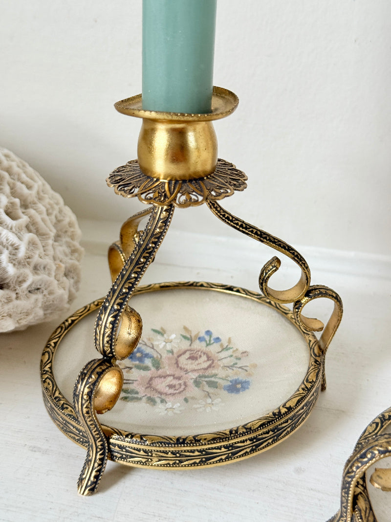 An ornate vintage-style candlestick holder with a gold and black filigree metal frame, a round glass dish with a pink floral print, and a light blue candle inserted, set against a white background.