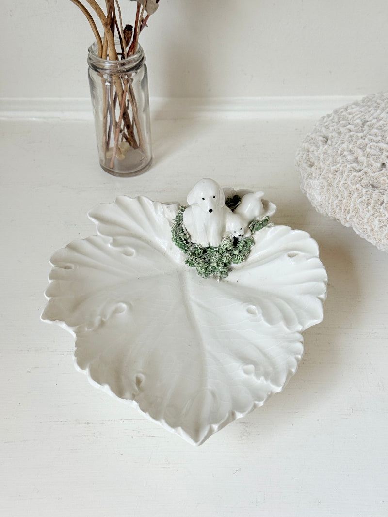 Italian ceramic dish featuring two white dogs on a sculpted leaf base, photographed on a white surface.