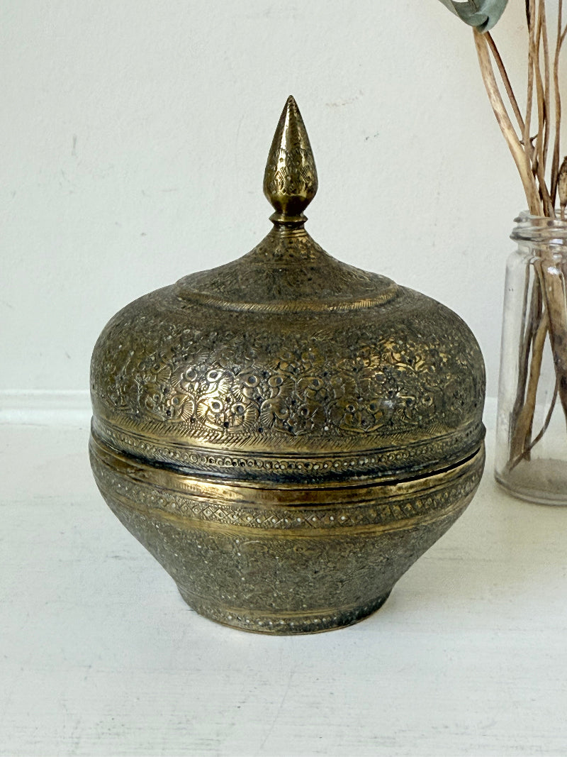 Indian Benares brass lidded jar with pointed finial and darkened interior, photographed on a white surface
