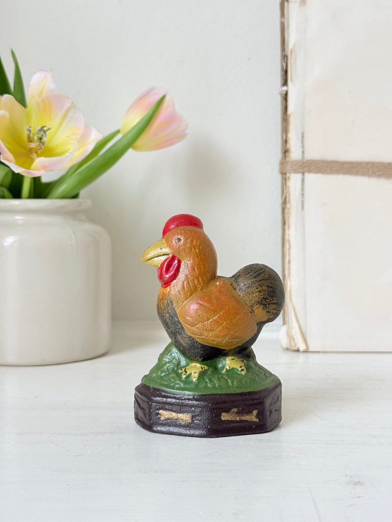 A small decorative rooster figurine with a red comb, yellow beak, orange-brown body, dark tail feathers, and a green base, displayed on a tabletop with a white vase of tulips and a neutral background.