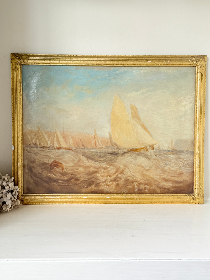 An oil painting of a sailing race on the sea, housed in an ornate gold frame, resting on a white shelf against a neutral wall.