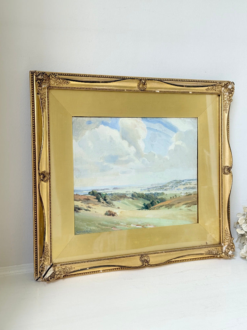 Framed oil painting of a rolling countryside landscape under a dramatic cloud-filled sky in an ornate gilt frame with wide mount.