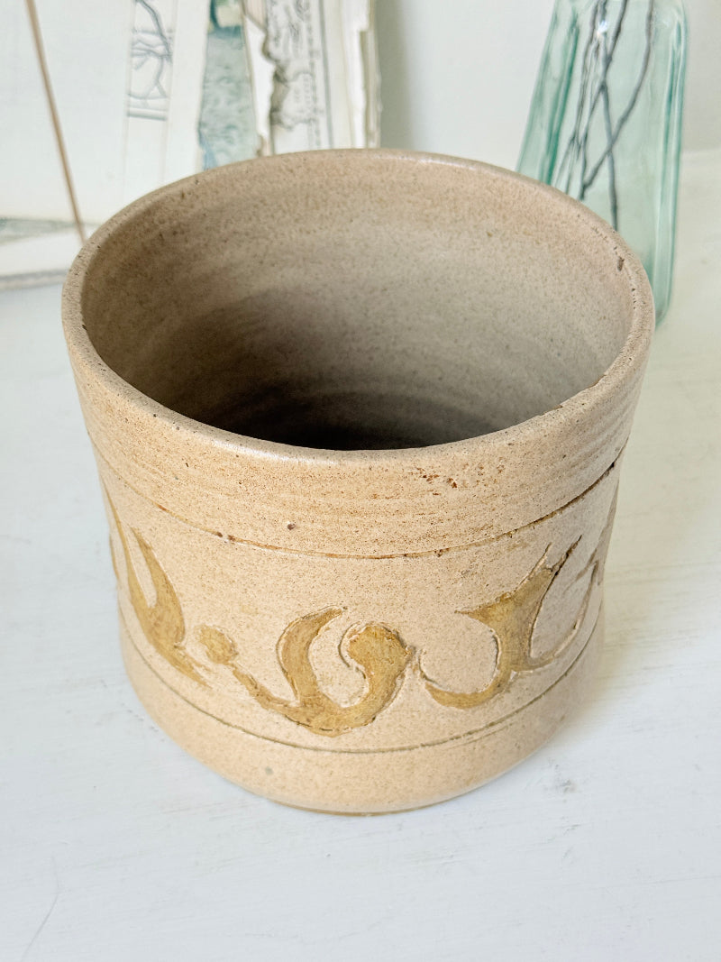 A handmade ceramic planter pot in a light beige stoneware finish, featuring a carved decorative band with organic gold-like leaf or scroll motifs around the lower half. The pot is photographed on a white surface with a softly blurred background containing glass bottles and framed art.
