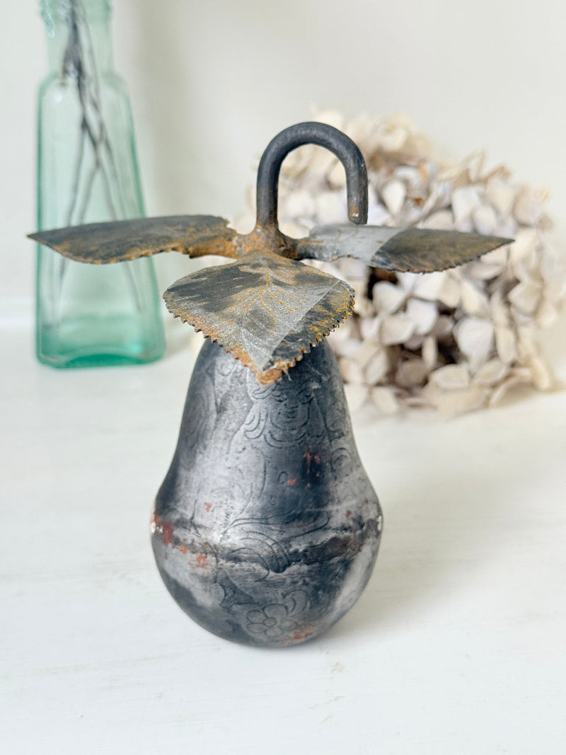 A vintage-style metal bird-shaped hanging ornament or decorative sculpture with a dark patinated surface, rusted brown highlights, and a curved top loop, photographed on a white surface with a blurred green glass bottle and dried flowers in the background.