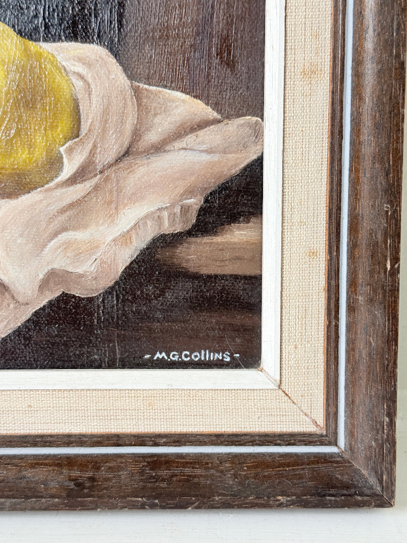 Close-up of a framed still life painting showing part of a yellow fruit, draped cream fabric, and a dark brown background, with the signature &