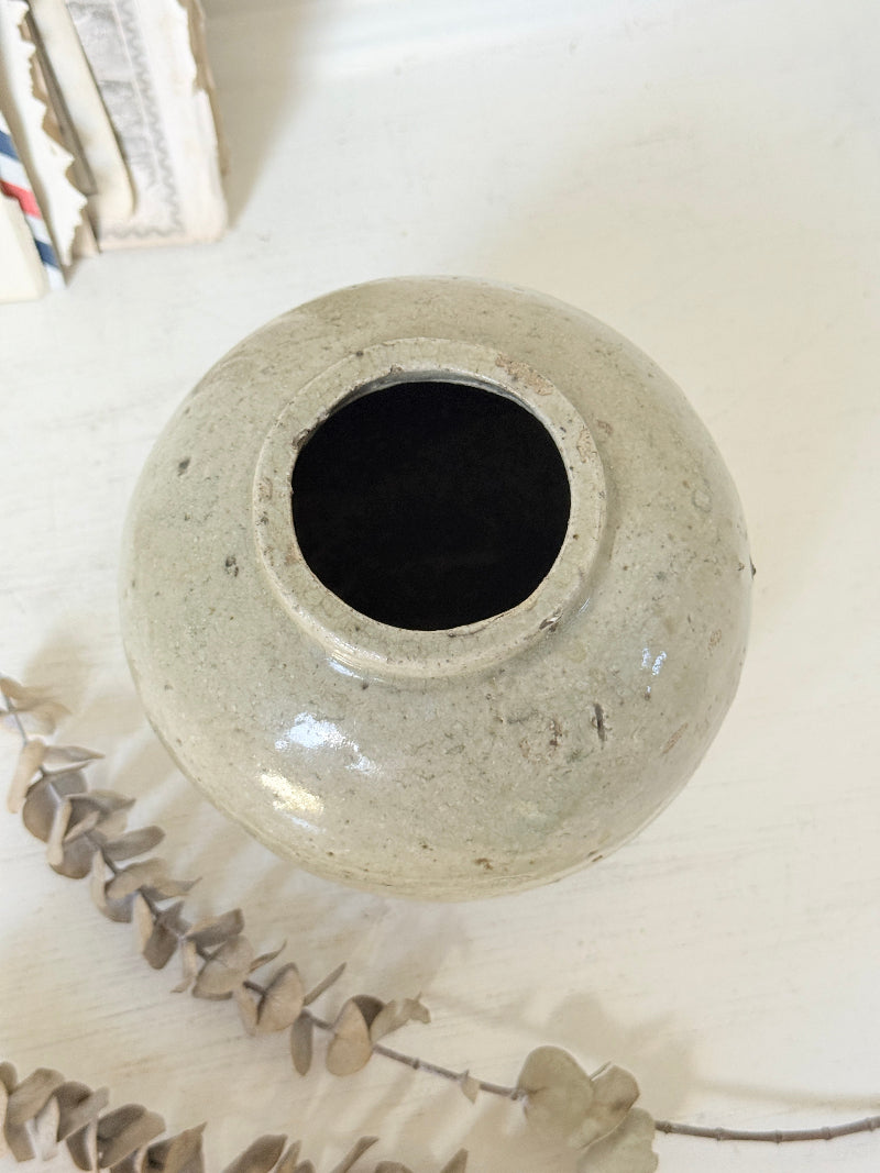 A small round ceramic vase with a wide opening, featuring a speckled light gray-green glaze and a dark interior, styled on a white surface with dried leaves in the foreground.
